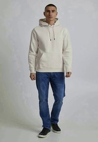 Light beige hoodie with a front pocket, drawstring hood, and ribbed cuffs, paired with blue jeans and black sneakers.