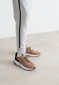 Καφέ αθλητικά παπούτσια με υφαντό άνω μέρος και μαύρες λεπτομέρειες, σε συνδυασμό με ανοιχτό γκρί φόρμες που έχουν μαύρη λωρίδα στο πλάι.