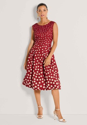 Frau trägt ein ärmelloses, knielanges rotes Kleid mit großen weißen Punkten, kombiniert mit weißen Sandalen mit hohen Absätzen und roten Ohrringen.