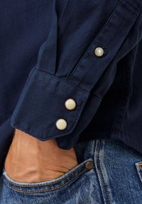 Polsino di una camicia a maniche lunghe blu navy con due bottoni rotondi di colore chiaro, caratterizzato da un tessuto testurizzato e dettagli stropicciati. Tasca dei jeans visibile.