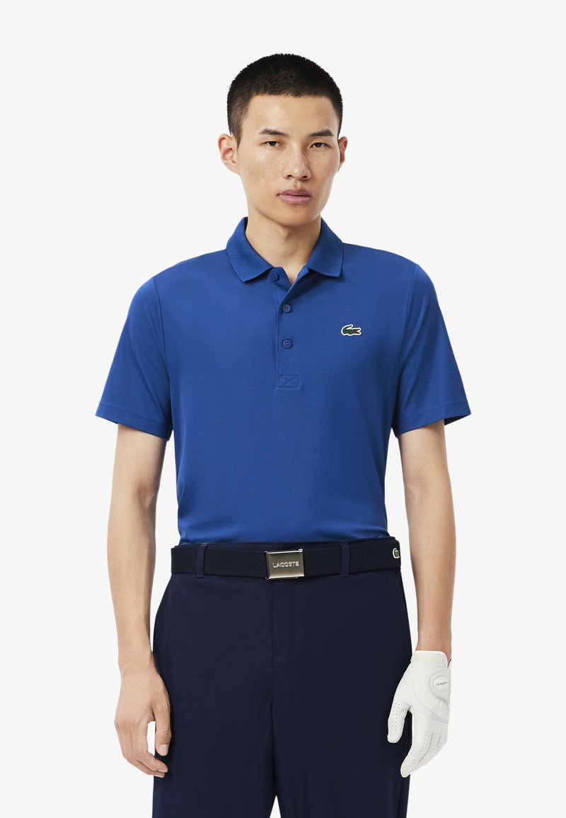 Blauwe polo met een zwart logo, korte mouwen en een knoopsluiting met drie knopen, gecombineerd met marineblauwe broek en een witte golfhandschoen.