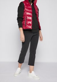 Rode puffer vest met horizontale stiksels, met rits zakken, gedragen over een zwart langemouwen top. Gekocht met cropped zwarte jeans en witte sneakers.