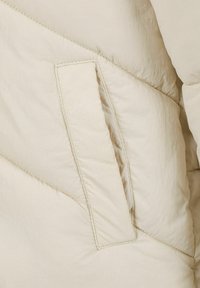 Beige puffjakke lavet af stof med et skråt syet design, med en firkantet klaplomme foran. Blød, let krøllet tekstur.