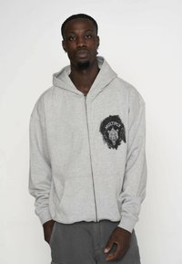Graue Zip-Hoodie mit einer Fronttasche, ausgestattet mit einem schwarzen Löwengrafik und dem Text "MULTIPLY" auf der Brust. Weiches Material, lockere Passform.
