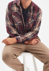 Geruite flanellen shirt in bordeaux, beige en marineblauw met twee borstzakken. Draagt over een zwart t-shirt, gecombineerd met beige broek.