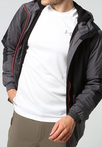 Giacca nera leggera con zip rossa a contrasto, dotata di cappuccio e polsini elastici, indossata sopra una maglietta bianca a maniche corte.