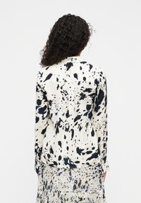 Πουκάμισο με κουμπιά σε κρεμ χρώμα με μαύρο μοτίβο πιτσιλιών, μακριά μανίκια και πιέτα στο πίσω μέρος. Το ύφασμα έχει λεία, ελαφριά υφή.