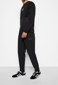 Svart sweatshirt med en kontrasterande svart panel och logotyp, tillsammans med svarta byxor med lila "adidas"-logotyp och svarta sneakers med vita accenter.