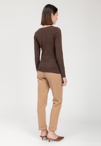 Braunes langärmliges Oberteil, taillierte Passform, kombiniert mit hellbraunen, schmal zulaufenden Hosen. Braune, offene Pantoletten mit spitzer Zehenpartie vervollständigen das Outfit.