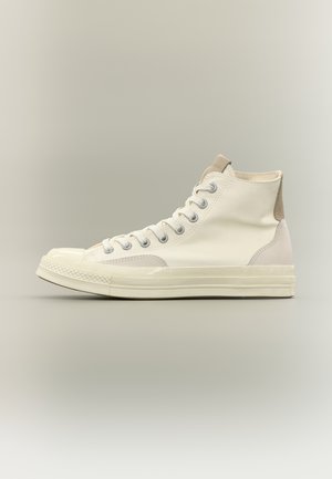 Balts augstās virsmas auduma sporta apavu modelis ar gumijas purngalu un zoli, metāla acīm un bēša zamšādas papēža plāksteri, fotografēts uz neitrālas fona.