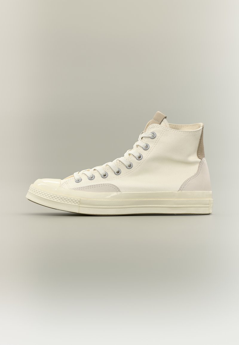 Λευκό πάνινο αθλητικό παπούτσι με ψηλό επάνω μέρος, καουτσούκ κάλυμμα στα δάχτυλα και σόλα, μεταλλικά οπές για κορδόνια και μπεζ σουέντ επιφάνεια στη φτέρνα, φωτογραφημένο σε ουδέτερο φόντο.