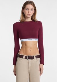 Bordeaux langemouw crop top met een witte Guess Jeans logo band. Paar beige high-waisted broeken met een bruine riem met een zilveren gesp.