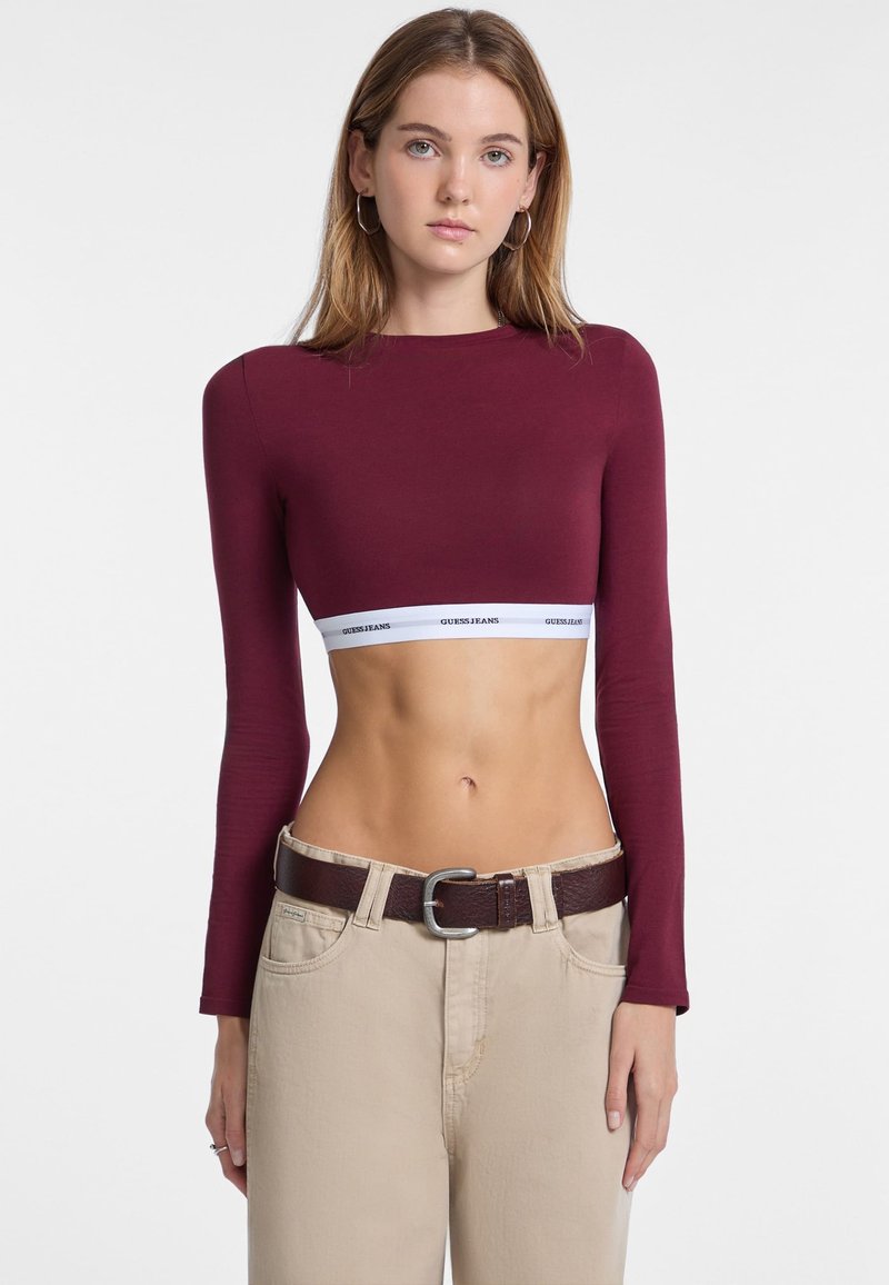 Bordeaux langemouw crop top met een witte Guess Jeans logo band. Paar beige high-waisted broeken met een bruine riem met een zilveren gesp.