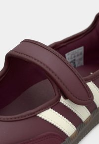 Burgundy athletic shoe with white stripes, featuring a wide velcro strap and a textured rubber sole. Smooth material and rounded shape.