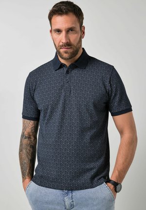Mand iført mørk mønstret poloshirt og lyseblå jeans, med tatoveret højre arm og sort armbåndsur på venstre håndled.
