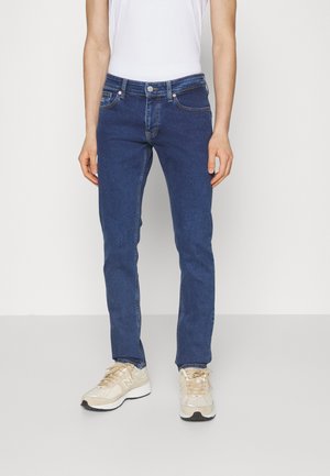 Persoon draagt strakke blauwe jeans en beige sneakers, staand tegen een effen lichte achtergrond.