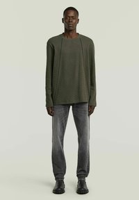 Pull en maille vert olive avec un motif texturé, associé à un jean gris. Le mannequin porte des bottines noires et se tient devant un fond vert clair.