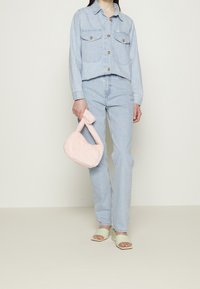 Jasnoniebieska kurtka jeansowa z dwiema kieszeniami na piersi, w zestawie z jasnoniebieskimi dżinsami. Trzyma miękką różową torebkę z futra sztucznego. Zielone sandały na obcasie z odkrytymi palcami.