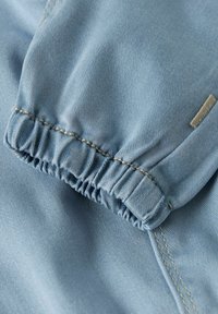 Lyseblått stoff med myk tekstur, med elastiske mansjetter og synlige sømmer. Lite logomerke på siden.