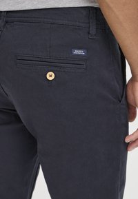 Gros plan d'une personne portant un pantalon chino bleu marine foncé avec une poche arrière boutonnée et une petite étiquette indiquant « Dedicated to Denim Perfection ».
