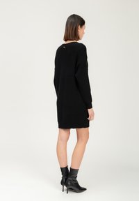 Vestito lavorato a maglia nero con maniche lunghe e vestibilità ampia. Presenta una trama a coste e un piccolo dettaglio dorato alla scollatura posteriore.