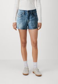 Shorts in denim azzurro chiaro, con orlo sfilacciato, design a cinque tasche e chiusura con bottone. Abbinati a calze bianche alla caviglia e sneakers chiare.