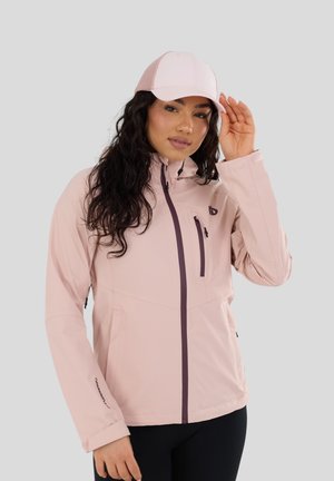 Donna che indossa una giacca leggera rosa chiaro con cerniera e un cappellino da baseball in rete rosa coordinato, che si aggiusta il cappellino con una mano, sfondo grigio neutro.