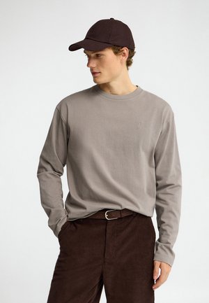 Jonge man die een taupekleurig shirt met lange mouwen draagt, donkerbruine ribfluwelen broek, een zwarte riem en een zwarte baseballpet, die naar de zijkant kijkt.