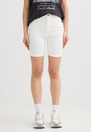 Persona que lleva shorts de mezclilla blancos, calcetines blancos, zapatillas plateadas y blancas, y una camiseta negra con estampado gráfico sobre un fondo liso.