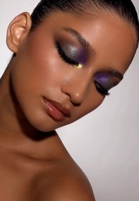 Nahaufnahme eines Gesichts mit metallisch lila und dunkelgrauem Lidschatten, glänzenden, nude Lippen, glatter Haut und nach hinten gekämmtem Haar.