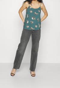 Top de tirantes finos con estampado floral, fondo verde azulado, flores rosas y blancas. Combinado con vaqueros grises de talle alto y sandalias negras de tacón.