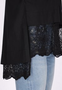 Blusa nera con orlo in pizzo a scallop, indossata sopra jeans blu chiaro. Il pizzo presenta motivi floreali intricati e una finitura testurizzata.
