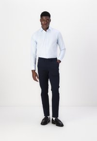 Light blue button-up shirt with long sleeves, dark navy slim-fit pants, and black loafers, standing against a white background.