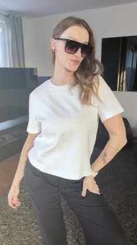 T-shirt en coton blanc à manches courtes porté avec un pantalon noir. Grandes lunettes de soleil noires avec des verres dégradés. Tatouages visibles sur les bras.