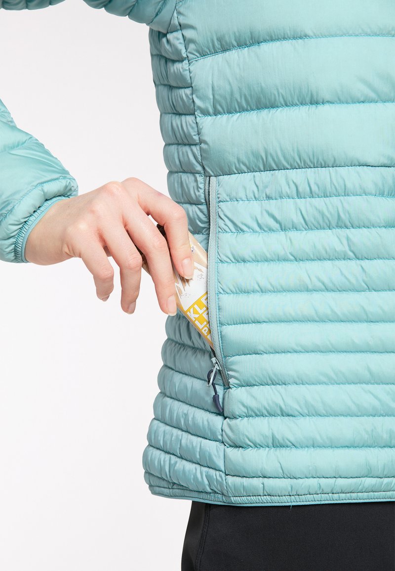 Chaqueta puffy azul clara con textura acolchada horizontal, que presenta un bolsillo lateral con cremallera, mano extendiéndose para sacar una tarjeta.