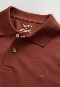 Tummanpunainen polo-paita, jossa on teksturoitu kangas, kaksi harmaata nappia ja brodeerattu logo rinnassa. Säännöllinen istuvuus -merkki näkyvissä.