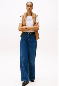 Beige gewatteerde vest over een witte t-shirt met tekst, gecombineerd met hoge taille blauwe wijde jeans en zwarte schoenen.