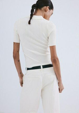 Žena s pletenom kosom, odjevena u bijeli rebrasti kratki rukav top i bijele hlače s crnim remenom, okrenuta leđima prema jednolikoj pozadini.