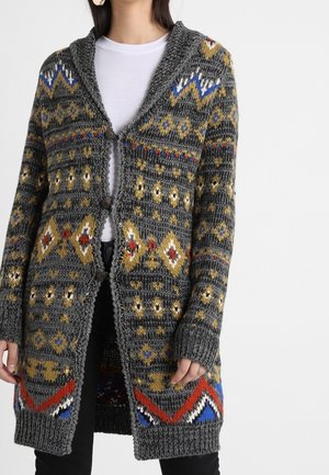 Vrouw draagt een lange, grijze gebreide cardigan met meerkleurige geometrische patronen en drie knopen, over een wit shirt en zwarte broek.