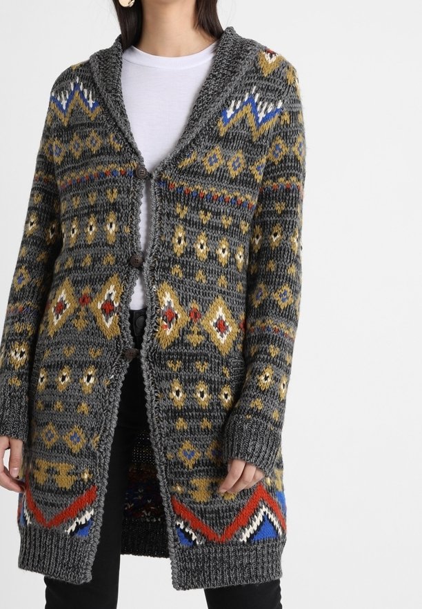 Femme portant un long cardigan gris tricoté avec des motifs géométriques multicolores et trois boutons, sur une chemise blanche et un pantalon noir.