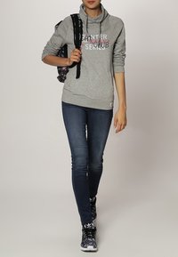 Felpa grigia con cappuccio e testo grafico, abbinata a jeans blu scuro e sneakers nere con motivo. Zaino nero con design floreale.