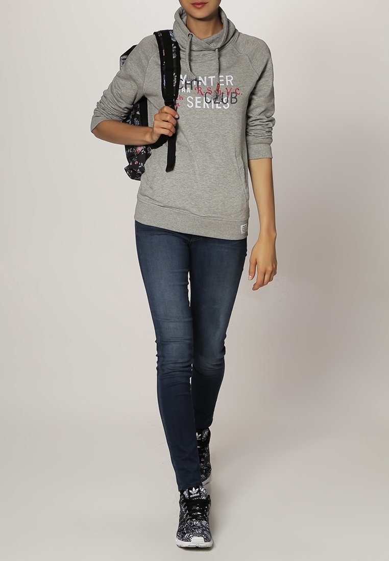 Felpa grigia con cappuccio e testo grafico, abbinata a jeans blu scuro e sneakers nere con motivo. Zaino nero con design floreale.