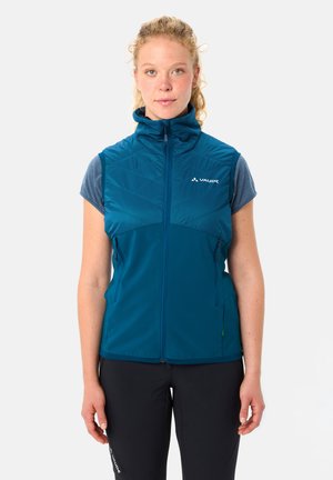 Frau trägt eine ärmellose, dunkelpetrolfarbene Weste mit Reißverschluss über einem blauen Kurzarmshirt und steht vor einem einfarbigen hellen Hintergrund.