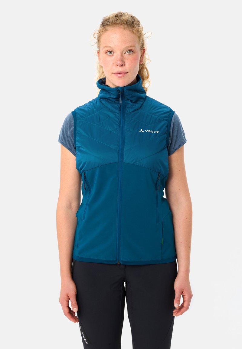 Frau trägt eine ärmellose, dunkelpetrolfarbene Weste mit Reißverschluss über einem blauen Kurzarmshirt und steht vor einem einfarbigen hellen Hintergrund.