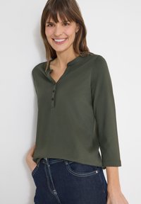 Femme souriante aux cheveux bruns, portant un haut vert olive à trois boutons et un jean bleu foncé, debout devant un fond blanc.