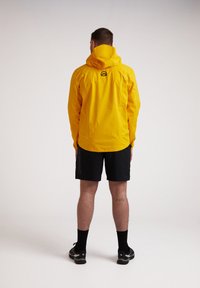 Gul vandtæt jakke med hætte, med et logo på ryggen. Båret over sorte sportsshorts, kombineret med sorte sokker og sko.