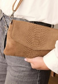 Bruine nepleren clutch met een gestructureerd slangenpatroon, voorzien van een klepsluiting en een polsbandje. Geplaatst tegen een lichtgekleurde shirt.