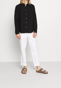 Černá košile z lnu s knoflíky, bílé kalhoty s ohrnutými manžetami a hnědé sandály s nastavitelnými popruhy na neutrálním pozadí.