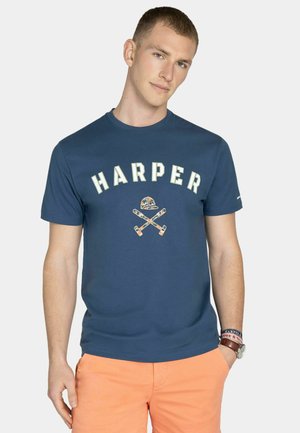Joven con camiseta azul marino "HARPER" con diseño de palos de golf cruzados y gorra, combinada con pantalones cortos de color naranja claro, manos en los bolsillos.