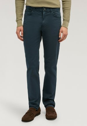 Mann trägt marineblaue Hose, braune Wildleder-Loafer und einen grünen, strukturierten Langarm-Pullover, steht vor einem schlichten Hintergrund.